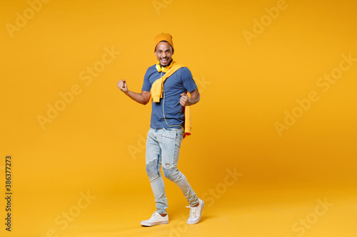Full length of happy joyful laughing young african american man 20s wearing basic blue t-shirt hat standing clenching fists doing winner gesture isolated on bright yellow background, studio portrait.