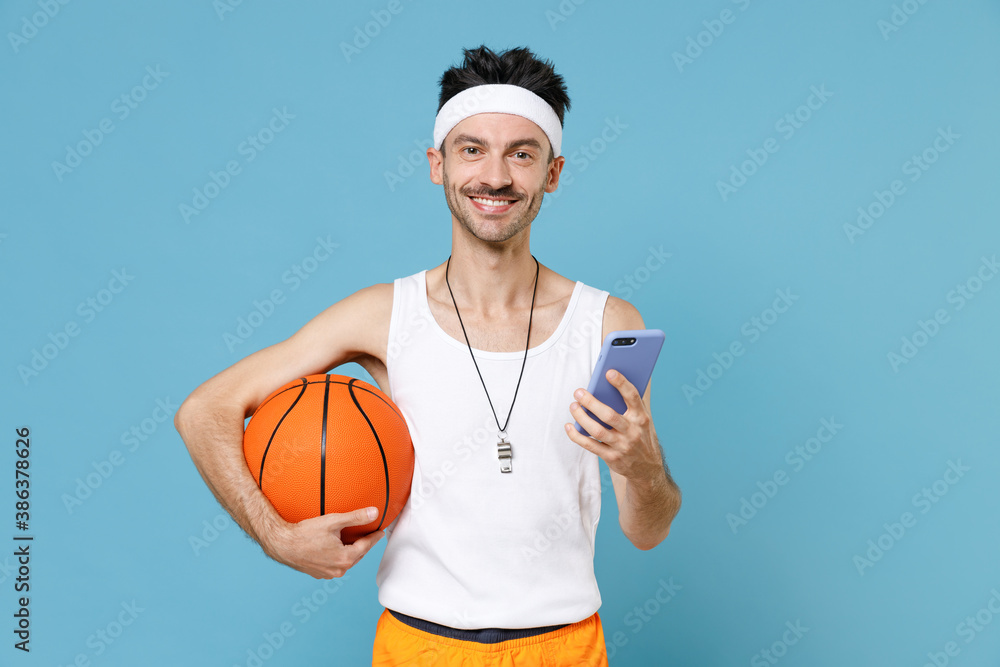 Smiling man basketball player with thin skinny body sportsman in headband shirt hold ball using mobile cell phone typing sms message isolated on blue background. Workout gym sport motivation concept.