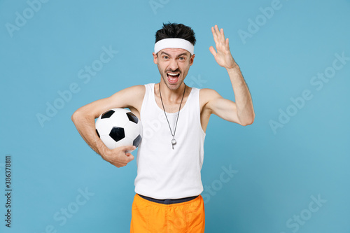 Angry sporty man football player with thin skinny body sportsman in headband shirt shorts whistle hold soccer ball screaming swearing isolated on blue background. Workout gym sport motivation concept. © ViDi Studio