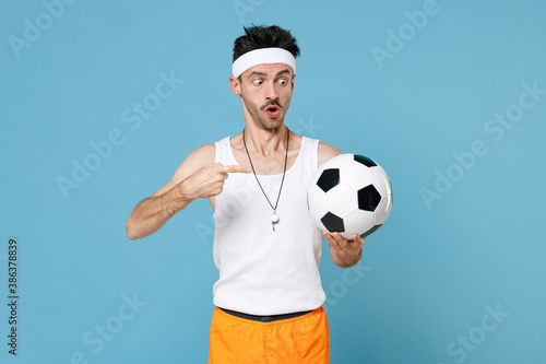 Shocked fitness man football player with thin skinny body sportsman in headband shirt shorts whistle pointing finger on soccer ball isolated on blue background. Workout gym sport motivation concept. © ViDi Studio