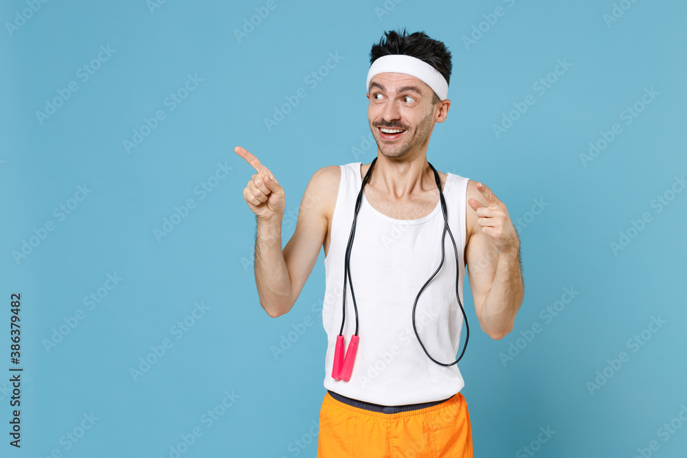 Excited young man with thin skinny body sportsman in white headband shirt shorts stand with skipping rope over neck pointing index fingers aside isolated on blue background. Workout gym sport concept. - obrazy, fototapety, plakaty 