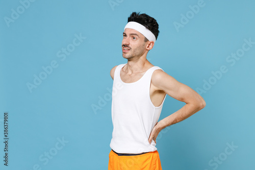 Dissatisfied young sporty fitness man with thin skinny body sportsman in white headband shirt shorts feels bad pain spasm back sprain isolated on blue background. Workout gym sport motivation concept. © ViDi Studio