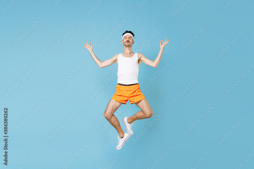 Full length young fitness man with skinny body sportsman in headband shirt shorts jumping hold hands in yoga gesture, relaxing meditating isolated on blue background. Workout sport motivation concept. Full length young fitness man with skinny body sportsman in headband shirt shorts jumping hold hands in yoga gesture, relaxing meditating isolated on blue background. Workout sport motivation concept.