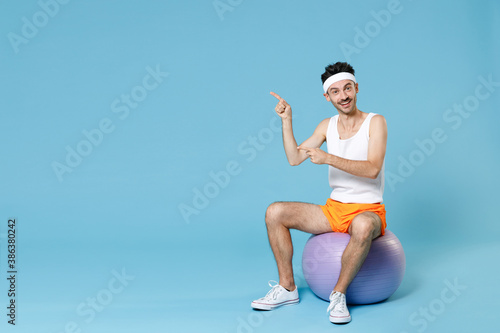Fototapeta Naklejka Na Ścianę i Meble -  Full length portrait funny young man with skinny body sportsman in headband shirt shorts sit on fitball pointing index fingers aside isolated on blue background. Workout gym sport motivation concept.