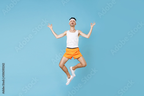 Full length young fitness man with skinny body sportsman in headband shirt shorts jumping hold hands in yoga gesture, relaxing meditating isolated on blue background. Workout sport motivation concept.