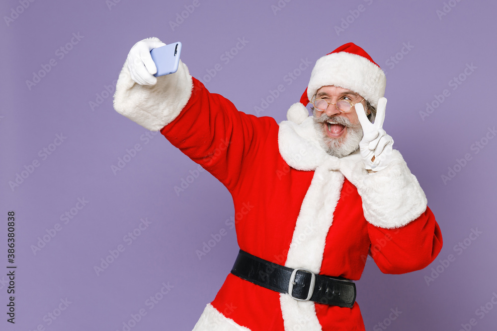 Blinking Santa Claus man in Christmas hat red suit coat glasses doing selfie shot on mobile phone showing victory sign isolated on violet background. Happy New Year celebration merry holiday concept. Blinking Santa Claus man in Christmas hat red suit coat glasses doing selfie shot on mobile phone showing victory sign isolated on violet background. Happy New Year celebration merry holiday concept.