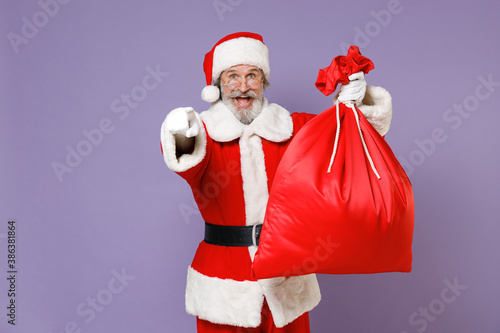 Excited Santa Claus man in Christmas hat red coat glasses hold bag with presents gifts pointing index finger on camera isolated on violet background. Happy New Year celebration merry holiday concept.