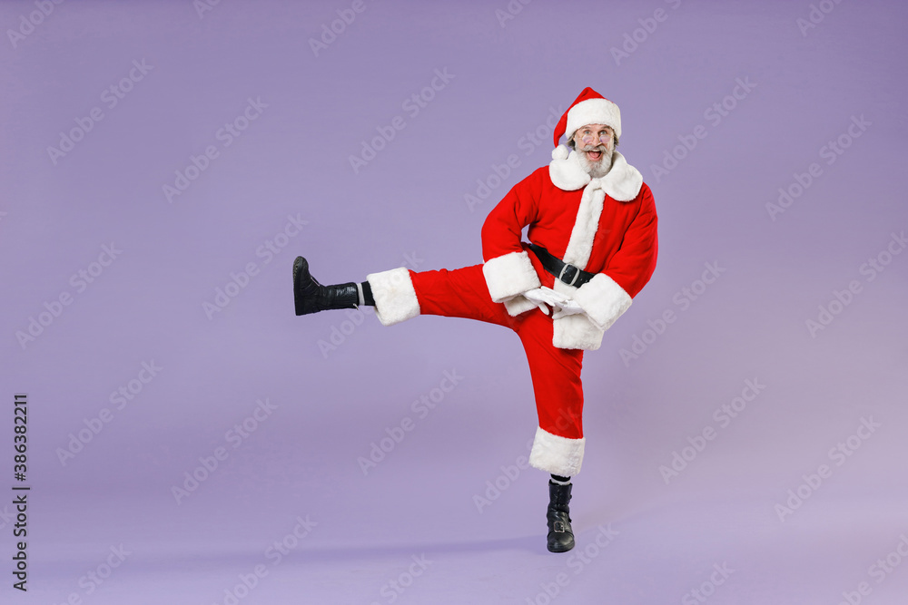 Full length portrait of excited Santa Claus man in Christmas hat red suit coat white gloves glasses dancing rising leg isolated on violet background. Happy New Year celebration merry holiday concept. Full length portrait of excited Santa Claus man in Christmas hat red suit coat white gloves glasses dancing rising leg isolated on violet background. Happy New Year celebration merry holiday concept.