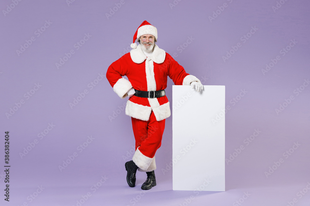 Full length portrait of smiling Santa Claus man in Christmas hat red suit coat glasses hold big white empty blank billboard isolated on violet background. Happy New Year celebration holiday concept. Full length portrait of smiling Santa Claus man in Christmas hat red suit coat glasses hold big white empty blank billboard isolated on violet background. Happy New Year celebration holiday concept.