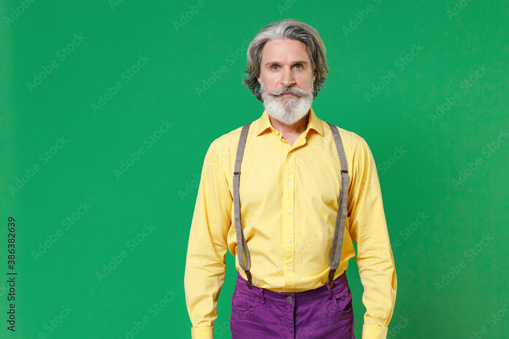 Handsome attractive elderly gray-haired mustache bearded man wearing casual basic yellow shirt suspenders standing looking camera isolated on bright green colour background, studio portrait.