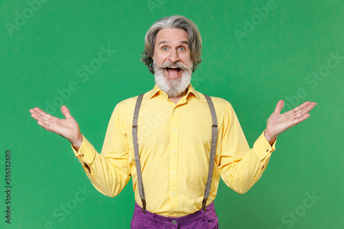 Excited surprised elderly gray-haired mustache bearded man in casual yellow shirt suspenders standing keeping mouth open spreading hands isolated on bright green colour background, studio portrait. © ViDi Studio