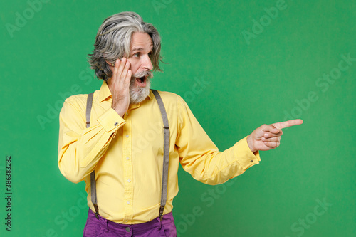 Shocked elderly gray-haired mustache bearded man wearing basic yellow shirt suspenders put hand on cheek pointing index finger aside on mock up copy space isolated on green background studio portrait. © ViDi Studio