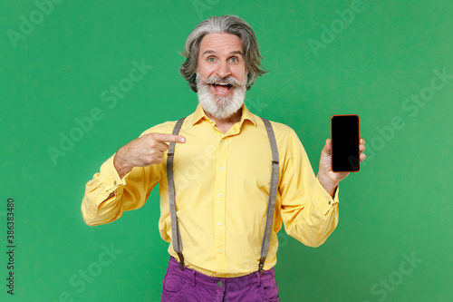 Shocked elderly gray-haired mustache man in yellow shirt suspenders pointing index finger on mobile cell phone with blank empty screen mock up copy space isolated on green background studio portrait. © ViDi Studio