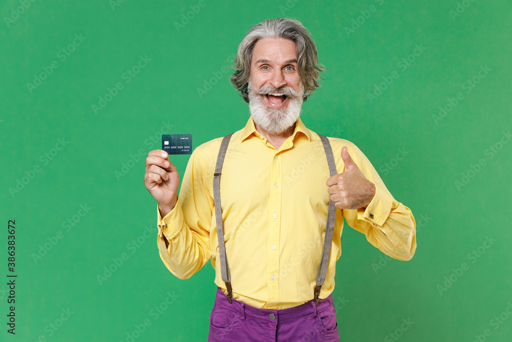 Excited elderly gray-haired mustache bearded man in casual yellow shirt suspenders hold credit bank card showing thumb up looking camera isolated on bright green colour background studio portrait.