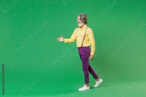 Full length side view of smiling elderly gray-haired mustache bearded man in yellow shirt suspenders standing with outstretched hand for greeting isolated on green colour background studio portrait. © ViDi Studio