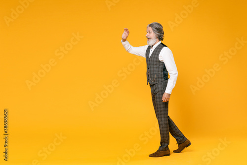 Full length side view elderly gray-haired business man in checkered suit waistcoat shirt waving greeting with hand as notices someone isolated on yellow background. Achievement career wealth concept.