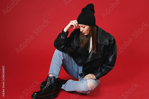 Full length of beautiful attractive young brunette woman 20s in casual black leather jacket white t-shirt hat sitting put hand on head looking down isolated on bright red background studio portrait.