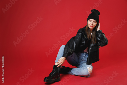 Full length of beautiful attractive young brunette woman 20s in casual black leather jacket white t-shirt hat sitting put hand on head looking camera isolated on bright red background studio portrait.
