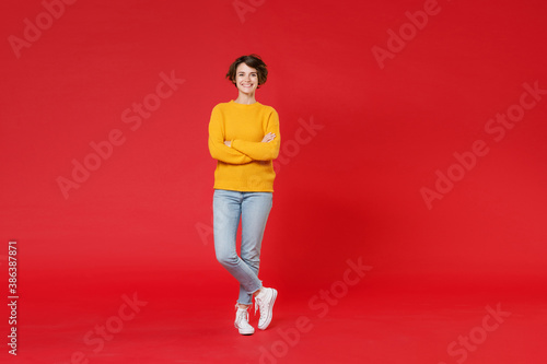 Full length of cheerful attractive young brunette woman 20s wearing basic casual yellow sweater standing holding hands crossed looking camera isolated on bright red colour background studio portrait.