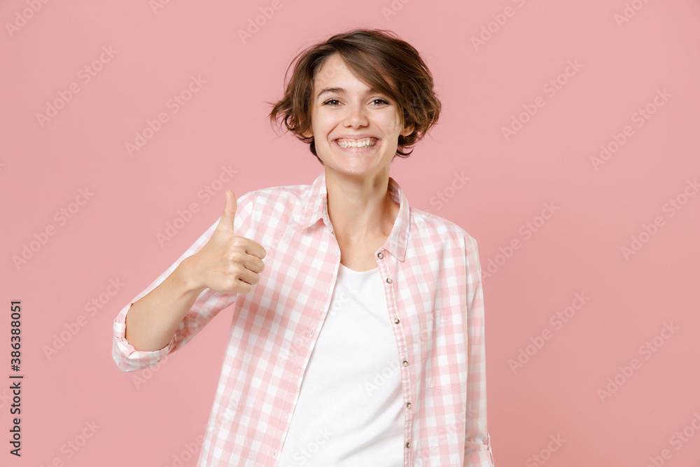 Smiling funny attractive beautiful young brunette woman 20s wearing casual basic checkered shirt standing showing thumb up looking camera isolated on pastel pink colour background, studio portrait. Smiling funny attractive beautiful young brunette woman 20s wearing casual basic checkered shirt standing showing thumb up looking camera isolated on pastel pink colour background, studio portrait.