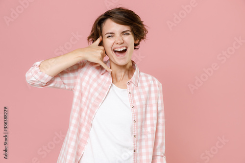 Laughing cheerful young brunette woman 20s wearing casual checkered shirt standing doing phone gesture like says call me back looking camera isolated on pastel pink colour background, studio portrait.