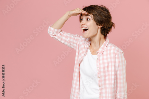 Excited surprised cheerful young brunette woman 20s in casual checkered shirt standing holding hand at forehead looking far away distance isolated on pastel pink colour background, studio portrait.