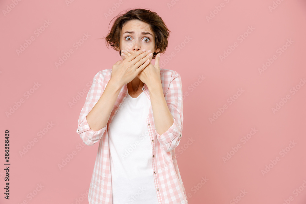 Shocked puzzled worried young brunette woman 20s wearing casual basic checkered shirt standing covering mouth with hands looking camera isolated on pastel pink colour background, studio portrait. Shocked puzzled worried young brunette woman 20s wearing casual basic checkered shirt standing covering mouth with hands looking camera isolated on pastel pink colour background, studio portrait.
