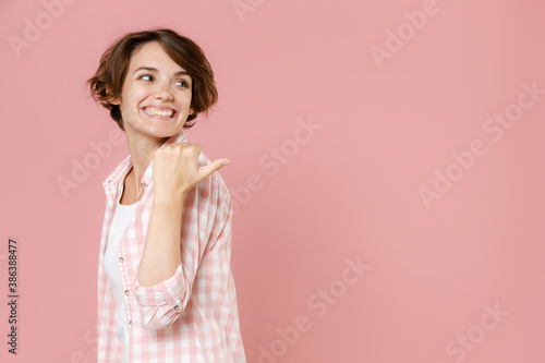 Side view of smiling pretty young brunette woman 20s wearing casual checkered shirt standing pointing thumb aside on mock up copy space isolated on pastel pink colour background, studio portrait.