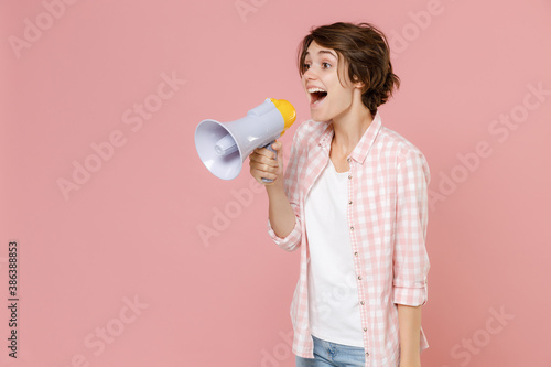 Excited shocked cheerful funny young brunette woman 20s wearing casual basic checkered shirt standing screaming in megaphone looking aside isolated on pastel pink colour background, studio portrait.