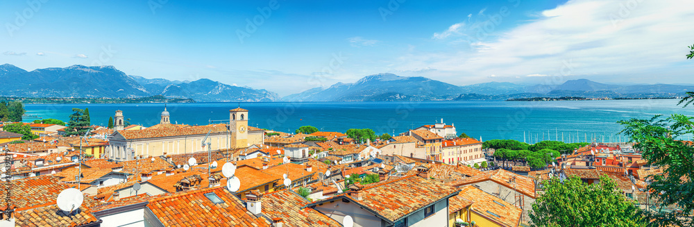 Panorama of Desenzano del Garda town with red tiled roof buildings, Garda Lake water, Monte Baldo mountain range, Sirmione peninsula, Lombardy, Northern Italy. Aerial panoramic view of Desenzano