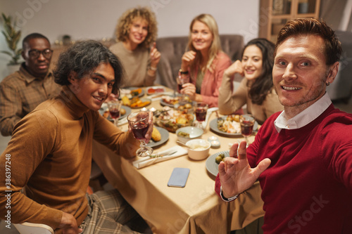 Portrait of smiling adult man looking at camera while taking selfie photo with friends and family at Thanksgiving dinner, copy space