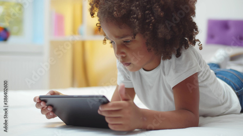 Adorable afro kid girl using digital tablet watching cartoons lying on bed at home