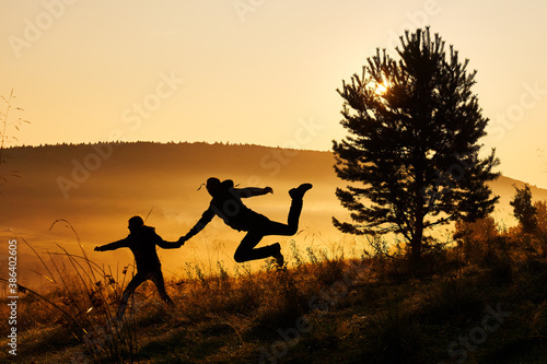 Silhouettes of a daughter and father making happy jump during beautiful golden sunrise. Foggy landscape in the mountains.