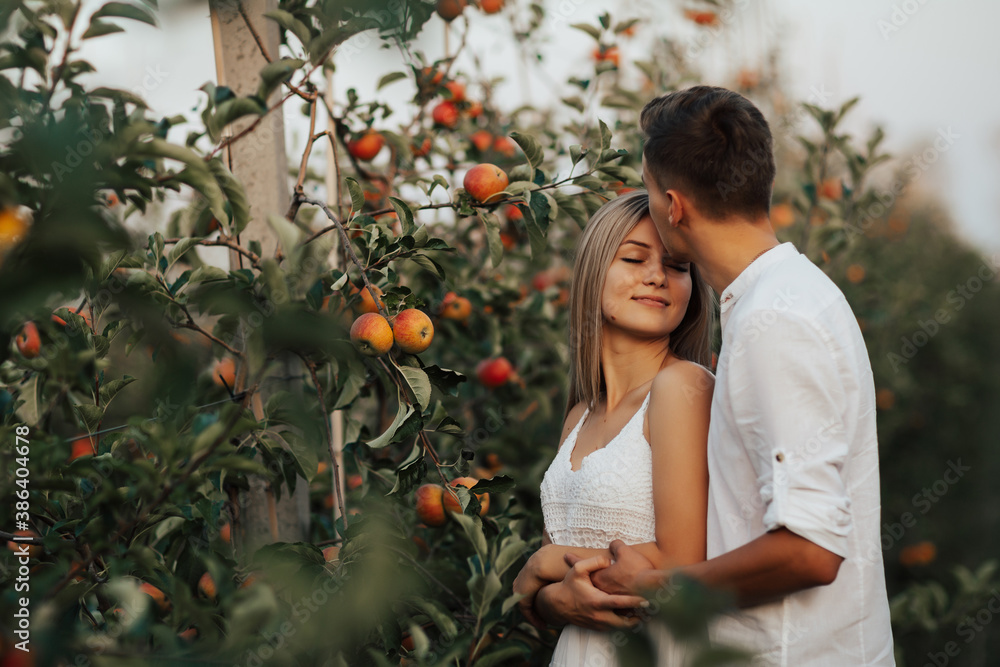 Happy bride and groom with closed eyes standing in embrace near trees in apple orchard. Man tenderly kissing him bride on the
forehead.