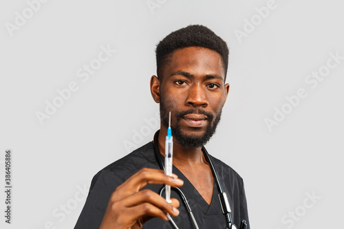 cute african doctor with stethoscope holds syringe in hand while standing against gray background. disease prevention and vaccination