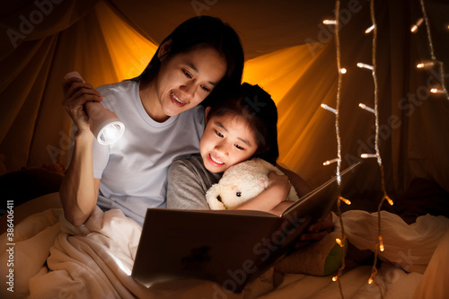 Family concept. Mother and child daughter reading book with flashlight together in children tent before bedtime. Happy mother read story book to her daughter in bed sheet tent.