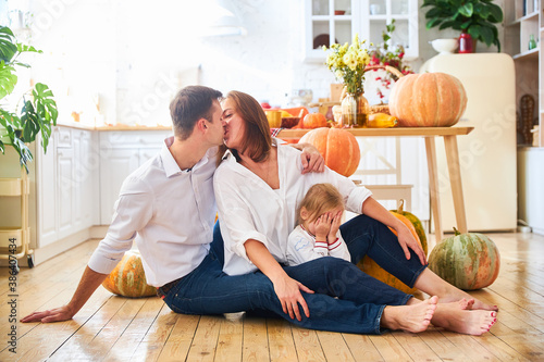 A happy family of three, mother, father and daughter, stand on a light kitchen, a wooden table with an autumn harvest - pumpkins, apples