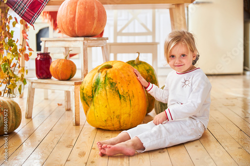 A little blonde girl sits on a wooden floor in a white kitchen next to a large orange autumn pumpkin