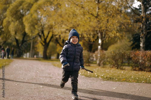 cute caucasian little boy running alone the lane in autumn park holding a stick in one hand and with his tongue stuck out © Yulia Raneva
