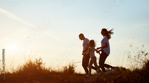 Young and beautiful family of three are jogging with their dog outside the city on the village road on the setting sun  side view  copy space