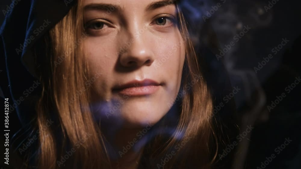 Close-up face of a young woman in a halloween hood extinguishes a candle blowing out and looking at the camera while flirting Close-up face of a young woman in a halloween hood extinguishes a candle blowing out and looking at the camera while flirting