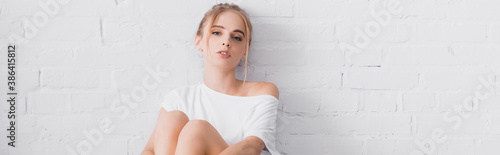 panoramic orientation of young blonde woman looking at camera while sitting near white wall