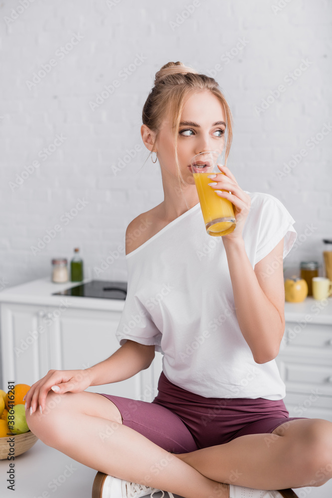 blonde woman sitting on kitchen table with crossed legs and drinking orange juice blonde woman sitting on kitchen table with crossed legs and drinking orange juice