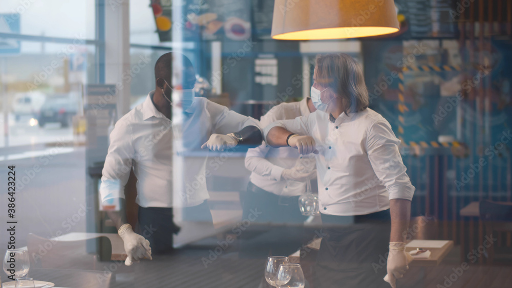 Obraz premium Diverse waiters in medical mask and gloves bumping elbows while setting tables in restaurant