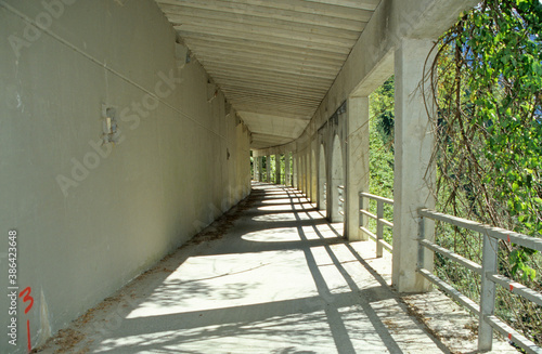 Long tunnel construction in mountain landscape. White concrete structure. Reinforced walls. Pillars and arches. Steel safety barriers. Sunlight and shadow patterns. Trees in background.