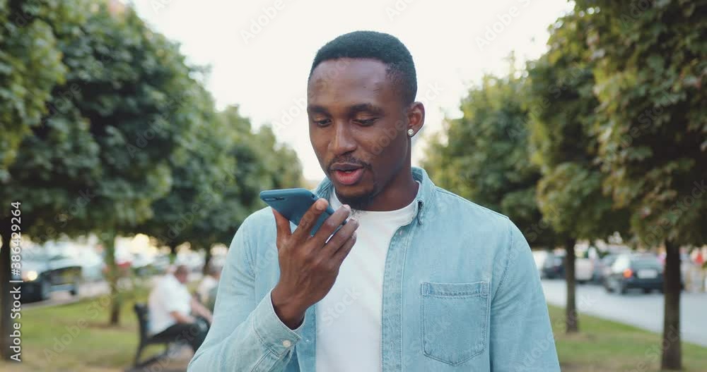 Handsome happy smiling young black-skinned man in casual clothes has video call while walking alone in the urban alley with green plantings in summer day Handsome happy smiling young black-skinned man in casual clothes has video call while walking alone in the urban alley with green plantings in summer day
