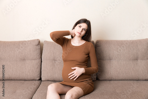 Young pregnant woman suffering from neck on sofa in home. Maternity healthcare concept