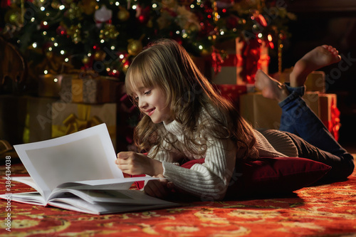 Happy young girl reading a story book in a cozy dark living room on Christmas eve. Celebrating Xmas at home. Christmas tree on the background.