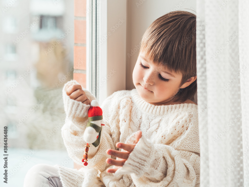 Fototapeta premium Kid with felt decorative snowman for Christmas tree. Boy in cable-knit oversized sweater. Cozy outfit for snuggle weather. Funny character with red Santa Claus hat. Winter holiday spirit. New year.