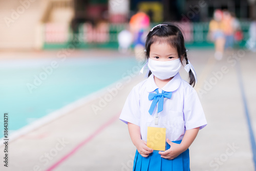 Child wearing white cloth face mask in school uniform is standing. Children go to school with new way of life (New normal). Cute kid looks at the camera. Kindergarten toddler age 3 years old.